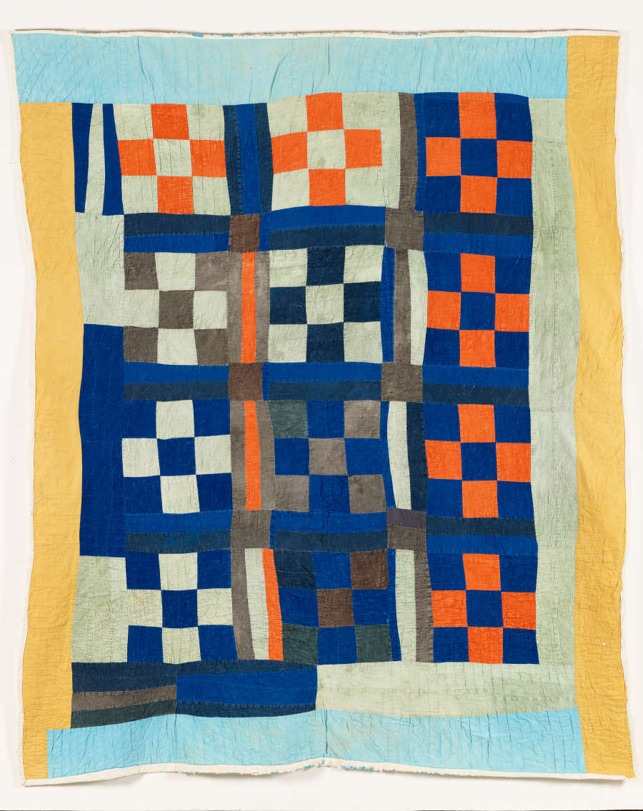 Quilt composed of dark blue, gray, orange, and light gray squares arranged in a checkered pattern. The squares are bordered and framed by uneven yellow, blue, and light gray strips. 