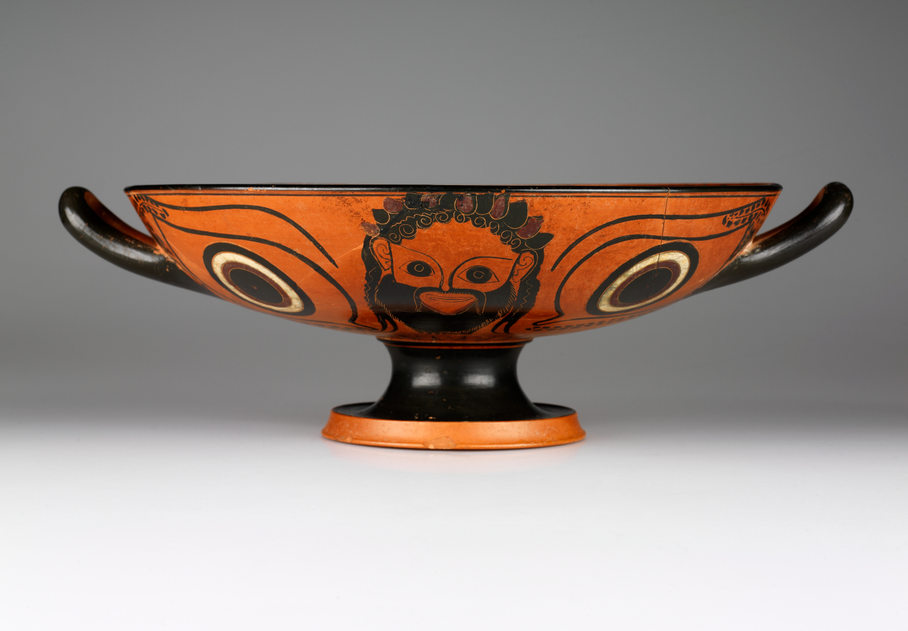 Terracotta and black cup with a wide mouth, short black neck, and orange base. One side features a bearded man flanked by two circular motifs rendered in black and white. 