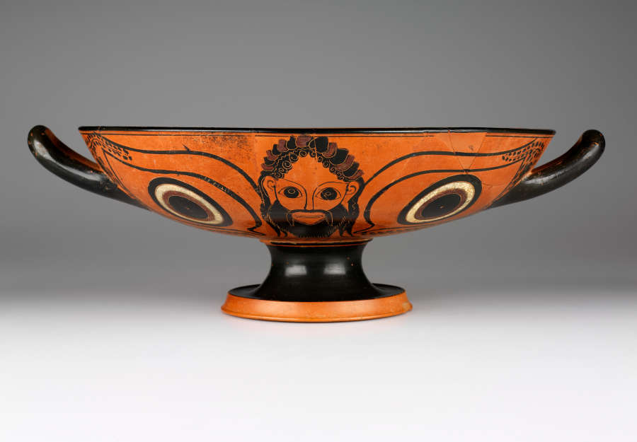 Terracotta and black cup with a wide mouth, short black neck, and orange base. One side features a bearded man flanked by two circular motifs rendered in black and white. 