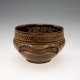 A earthenware bowl with a rounded form, lower half painted dark brown, upper half decorated with black and brown geometric patterns, and featuring a raised, undulated handle facing the viewer. 