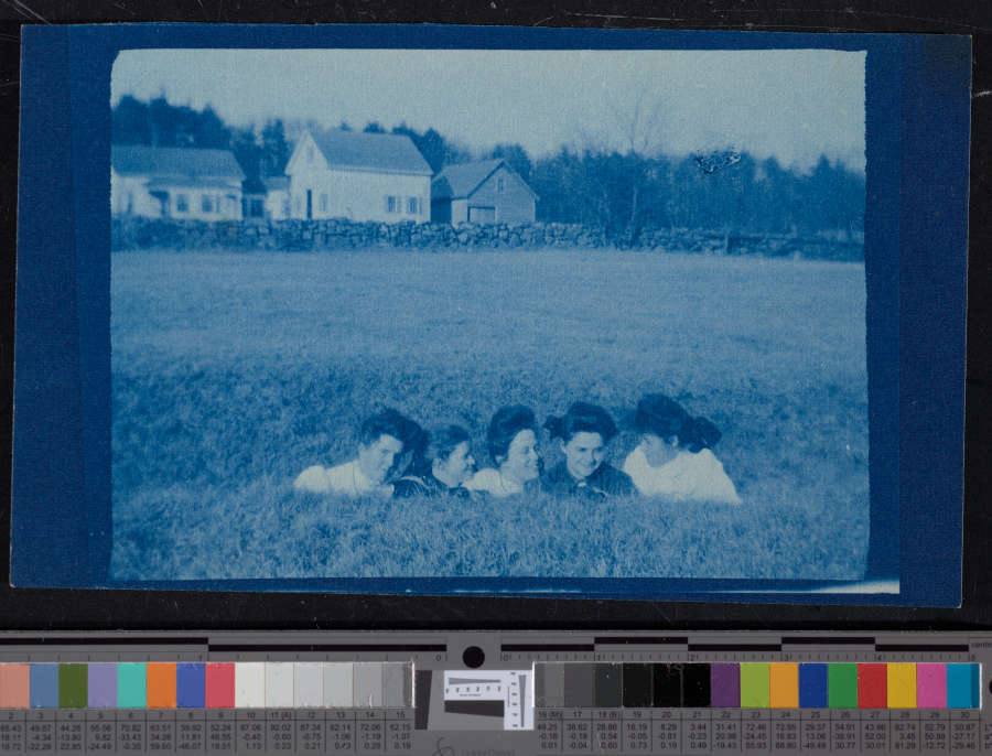 Blue-toned vintage snapshot of five smiling young light-skinned women. They are in the middle of a field, and only their heads and shoulders are visible. White houses rise behind them. 