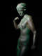 Cropped image of a gray metal sculpture with a green patina of a standing crowned nude woman, one arm down, grasping an object, and the other curled towards her shoulder.