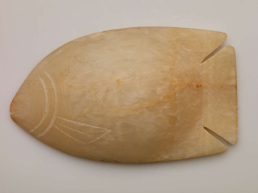 Bottom of an ovular tan dish shaped as a fish. The left side of the bowl forms the head, with engraving on the surface highlighting the fish’s face and fins.