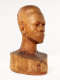 Three quarter front view of a glossy wooden bust depicting the head of a male figure with afrocentric facial features. He has a stern expression and an intense gaze.