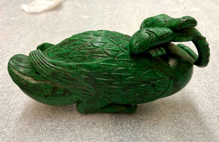 Green stone sculpture of a seated duck in profile, its legs tucked under its body and its mouth holding a fish, looking towards the viewer. The duck’s feathers resemble leaves. 