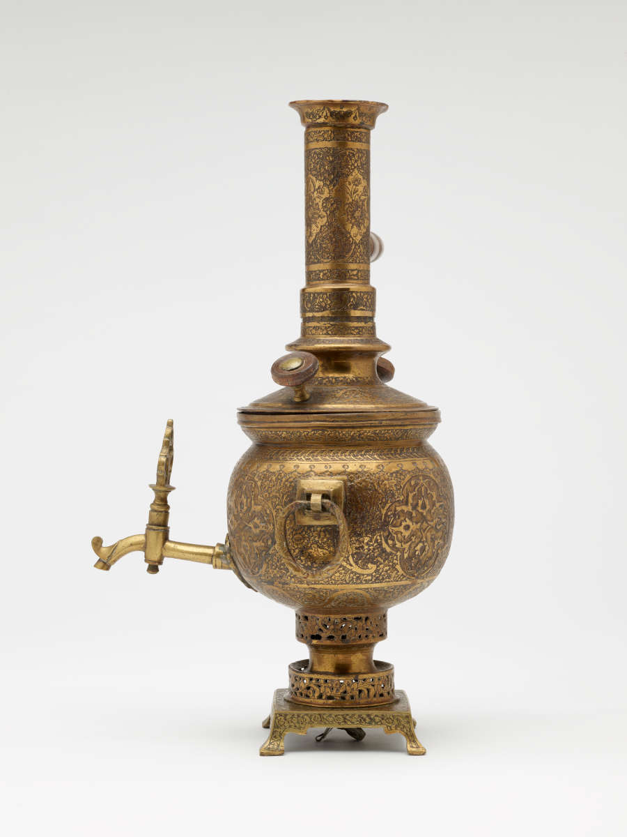 A brass and wood urn that has a decorative foot, lid, various handles and rings, and spout. The entire vessel has highly decorative engravings.