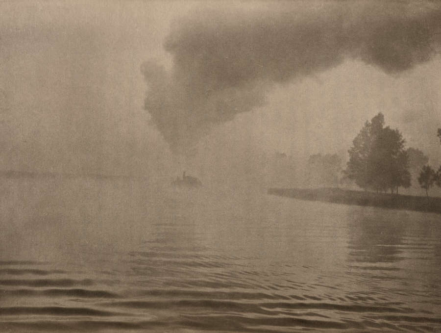 A hazy black and white image depicting a calm body of water with trees coming from the right side and a cloud of smoke rising from the background.