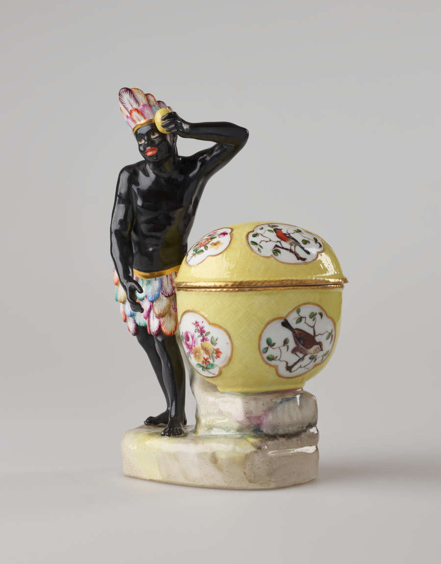 A sculptural figure with black skin, a colorful headdress and skirt. This figure is standing next to a bright yellow covered sugar basket with floral decorations.