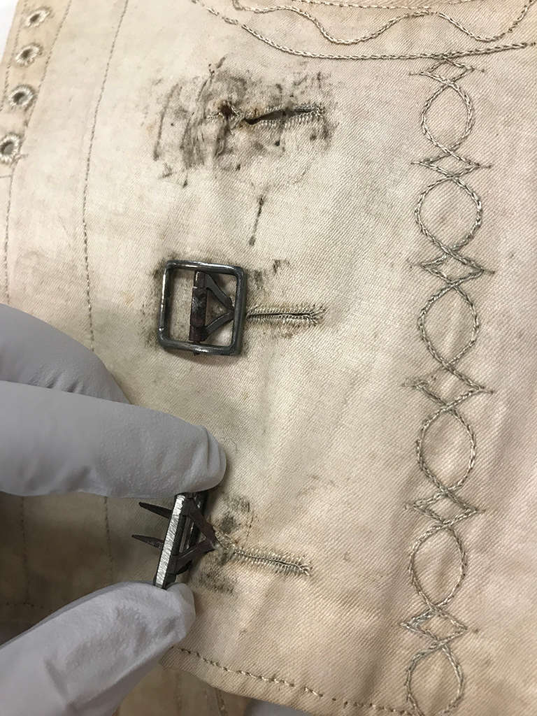 A gloved hand holds a small metal buckle near an aged, cream-colored fabric with worn embroidery and strap slits. 
