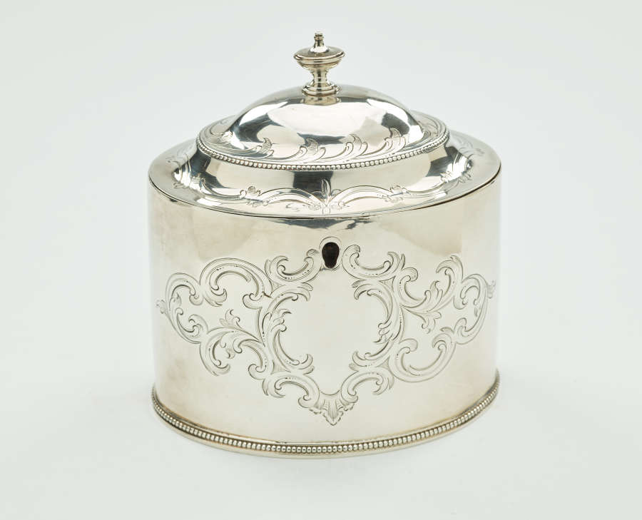 A silver tea caddy with a lid and sculptural finial. There are swirling engravings along the body and lid.