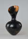 An asymmetrical, hourglass-shaped black vessel with a reflective finish. Its mouth slopes downward toward the viewer, exposing a bright terracotta interior.