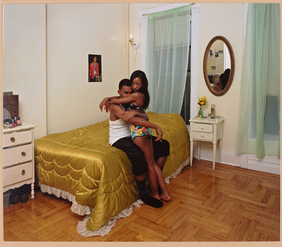 Photo of a dark-skinned young couple sitting on a bed, embracing. She wears shorts and a tank, he wears an undershirt and jeans. The bedspread is gold satin with fringe. 