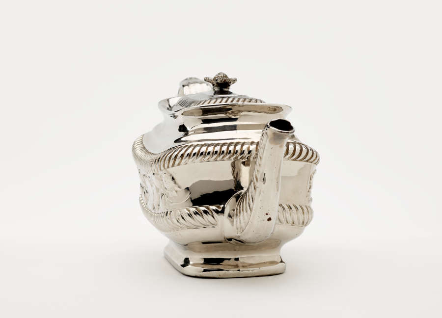 A silver teapot with a decorative handle, a rounded square body and spout with sculptural decorations.