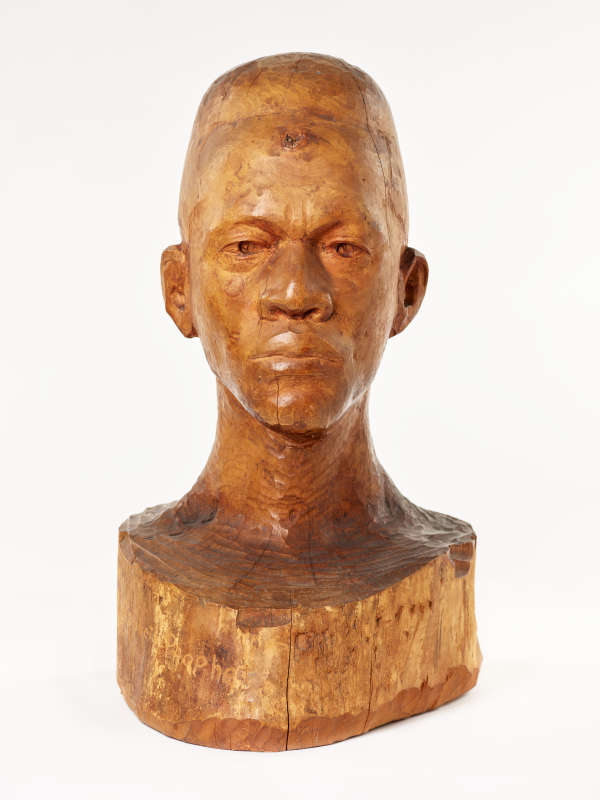 Front view of a glossy, carved wooden bust depicting the head of a male figure with afrocentric facial features. He has a stern expression and an intense gaze.