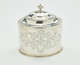 A silver tea caddy with a lid and sculptural finial. There are swirling engravings along the body and lid.