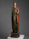 A tall, weathered wooden statue of a robed figure with a serene expression, draped in green and red garments, standing upon an octagonal base.