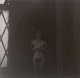 A dimly lit photograph of a nude figure holding a camera standing by a hatched window in soft light.
