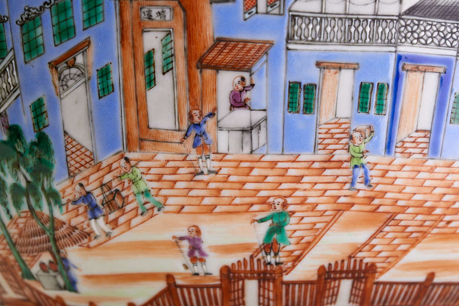 A close up of a painted ceramic depicting figures walking and interacting on an orange brick street, bordered by blue buildings with green windows and ornate wooden fencing.