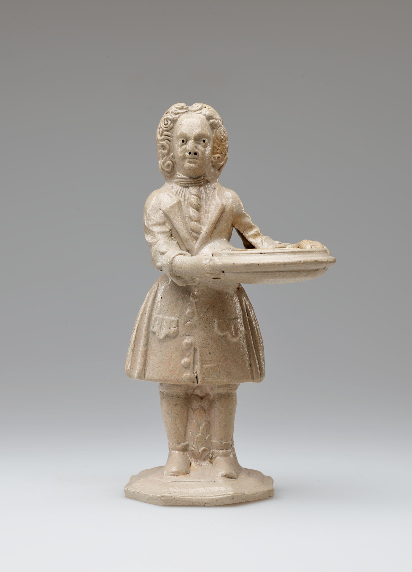 A cream colored sculptural figure holding a tray, dressed in historical clothing with tightly curled hair.
