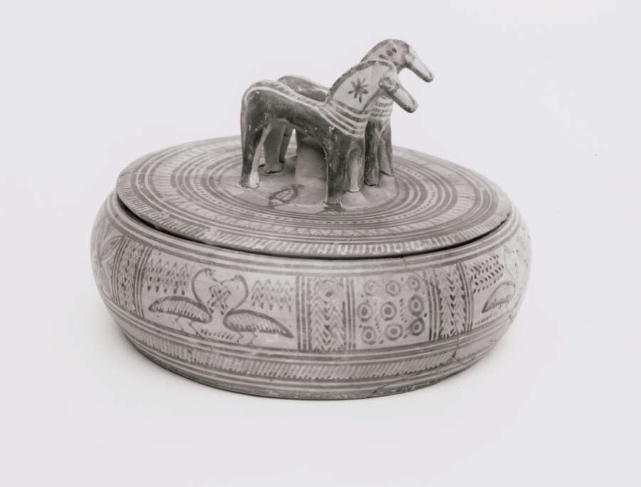Black and white side-view of a short and wide round lidded box, decorated with patterns and swan illustrations. The lid’s knob is in the shape of two standing horses. 