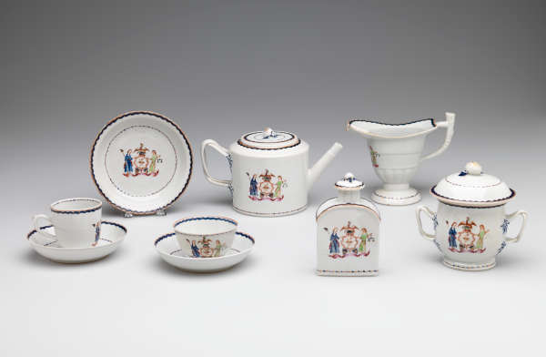 White porcelain tea set with blue and yellow trim, each piece featuring a painted emblem of an eagle, wreath, and two figures. Set includes a teapot, bowl, cups, and pitcher.