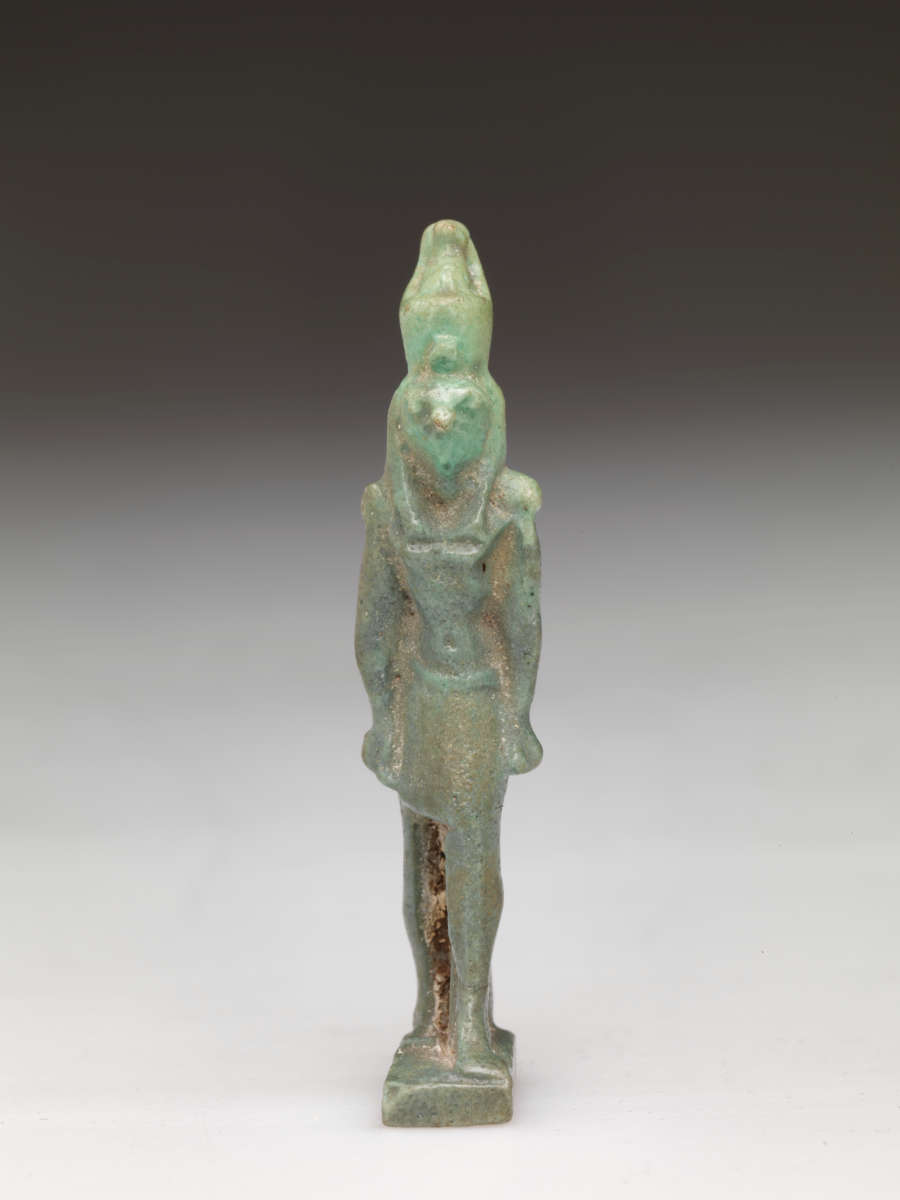 Standing light-jade and brown sculpture of a standing person with a tall head, narrow torso, and straight arms. The left leg has brown specks on it while the headdress is light and translucent.