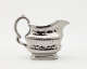  A silver creamer with a decorative handle and a rounded square body with sculptural decorations.
