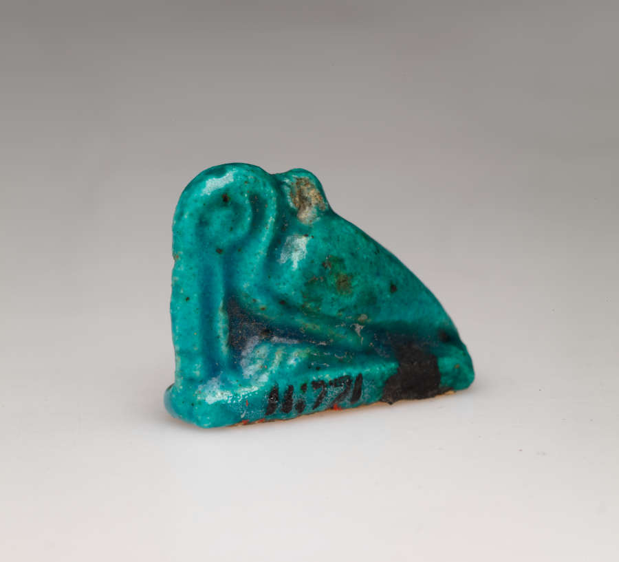 Turquoise amulet at an angle, of a crouching pelican-like bird with a long beak pointed downwards, head tucked into its neck, where there is a small loop. It is labeled in black ‘11.771’.