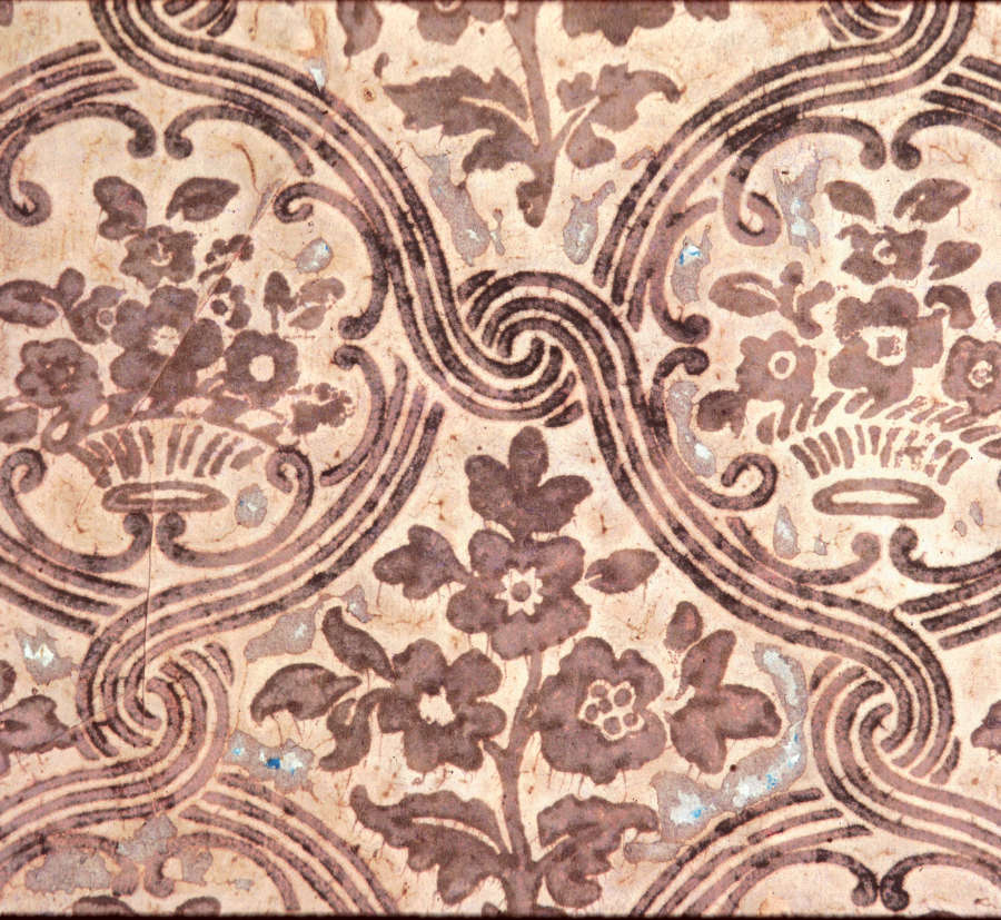 Close up segment of vintage wallpaper depicting a rustic pattern of brown florals and intricate swirl motifs on a beige, textured background.