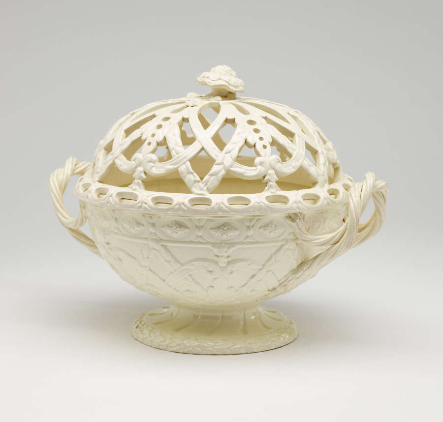A cream-colored basket with sculptural woven handles, sculpted foot, and a lid with cutout designs.