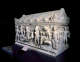 A marble coffin with an angled roof and deep-set carvings of human figures engaging in a narrative, displayed in a dark gallery. 