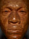 Close-up front view of an wood-carved face in high-contrast lighting depicting a male figure with afrocentric facial features. The sculpture fills mostly the whole frame, highlighting the man’s stern expression.