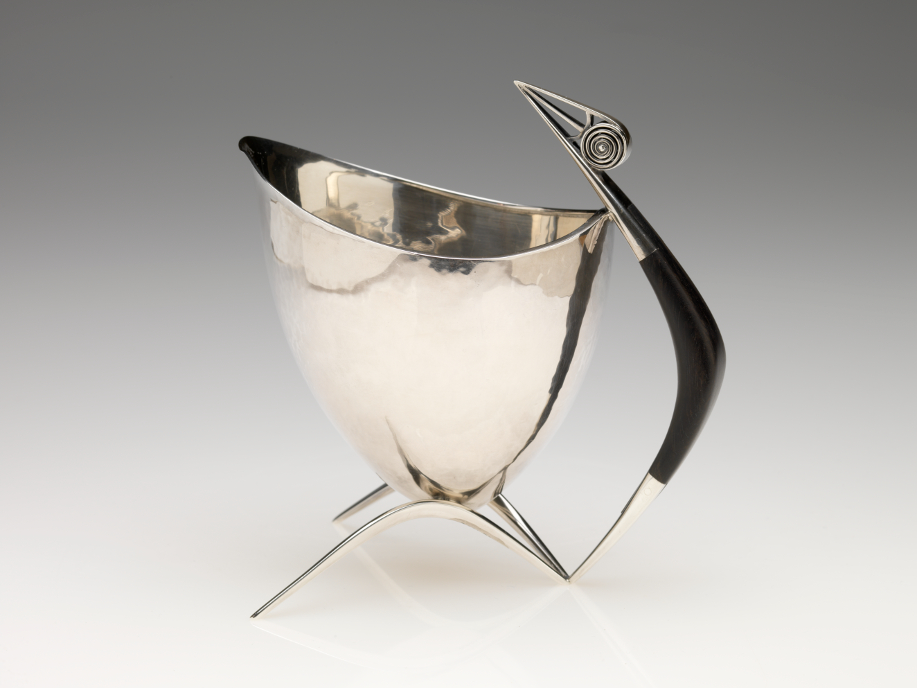 Sleek silver vessel with a curved body atop three pointed legs, featuring a black handle with a spiral detail and sharp ends which stretch above the vessel's top.