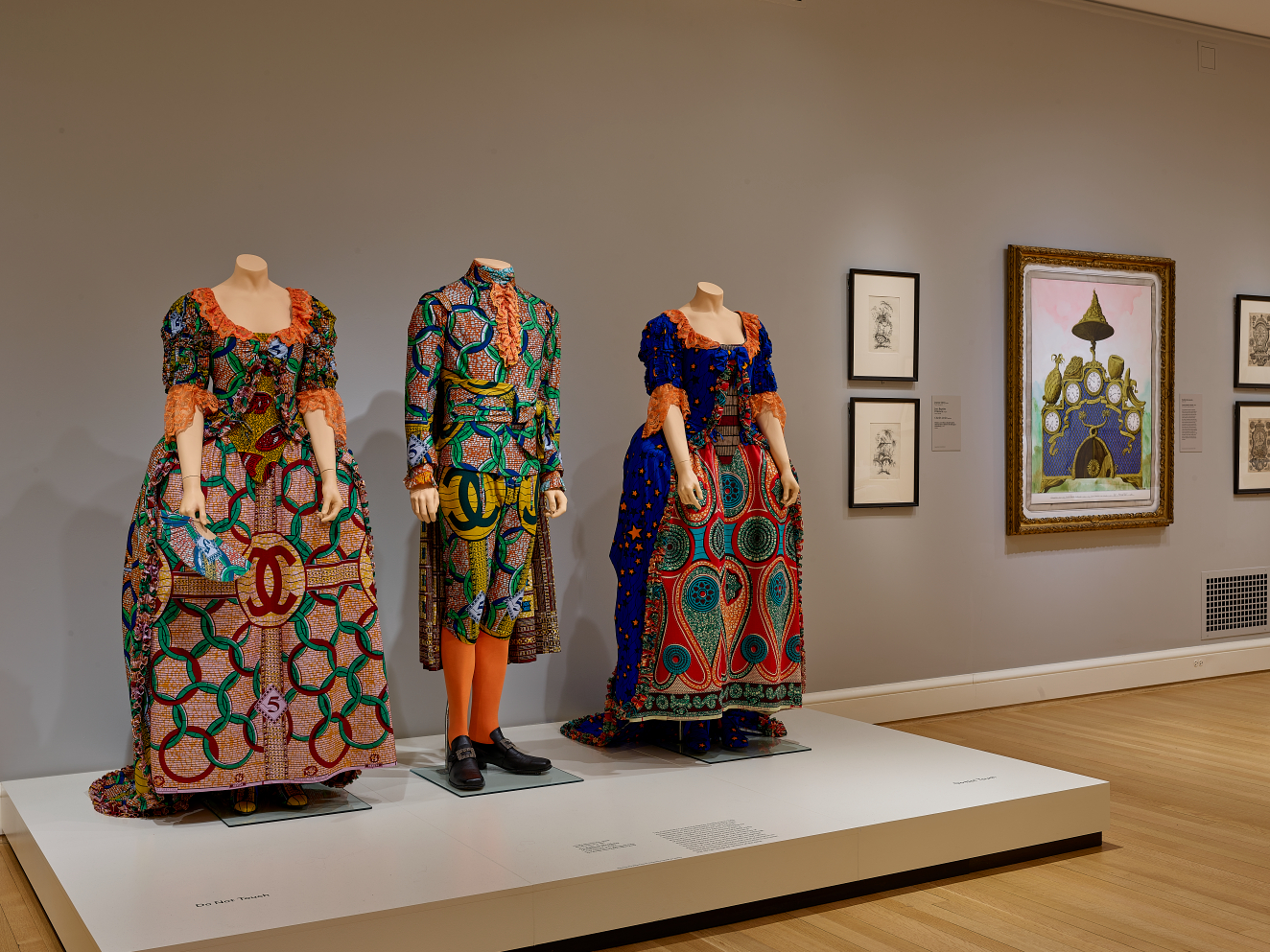 Three pale mannequins wearing brightly patterned Victorian clothing in varying shades of orange and blue stand to the left of small framed drawings and a larger colorful illustration.