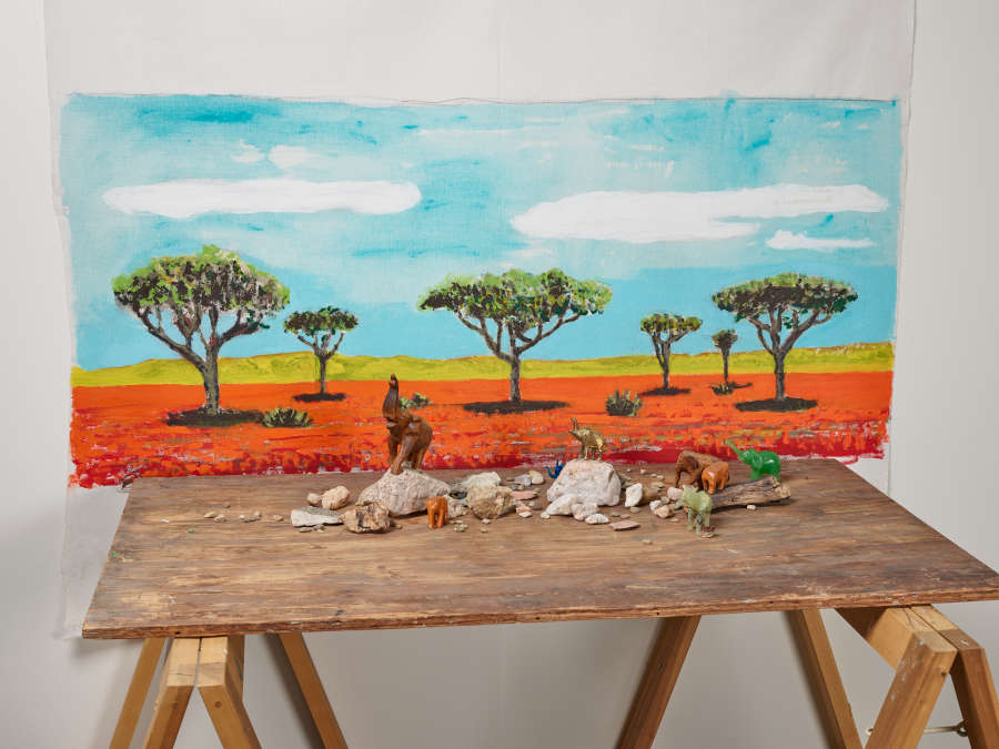 Painting of trees in orange soil against a blue cloudy sky is hung behind a wooden table rocks and toy animal figures on a table in front.