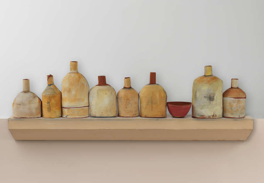 Assortment of ten ceramic vessels in various shapes, glazed in muted tans, ochres, and light greys with one deep red bowl, arranged on a beige shelf against a plain wall.