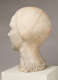 Three quarter back view of a marble sculpture depicting the head of a figure. The head features a tight hairstyle and delicate facial details. A signature is carved at the base.