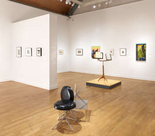 Several 2D works on white walls. Two 3D works are on the wood floor. One includes two barstools, the other is a music stand displaying materials.