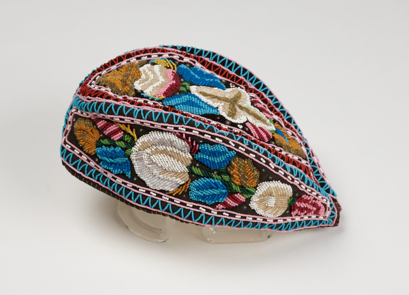 An almond shaped embroidered cap, which features 3 blocks of blue, white, green, and pink floral embroidery which are framed and divided by blue, pink, and white geometric patterns.