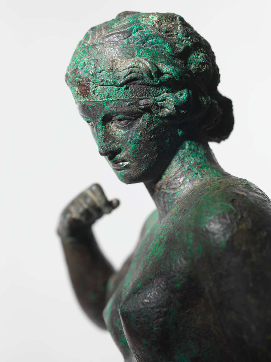 Close-up of a gray metal sculpture with a green patina, of a crowned woman looking to one side with one arm curled towards her shoulder. The photo is cropped mid-torso.