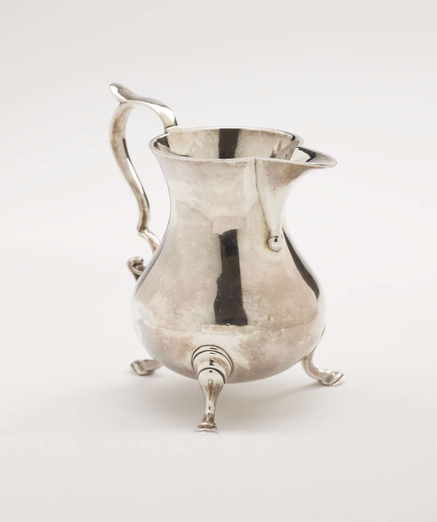 A silver creamer with a sculptural handle and three protruding feet.