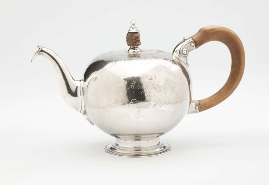 A bulbous silver teapot with a wooden finial and handle connected with decorative silver pieces. There are delicate engraved marks on the body and lid.