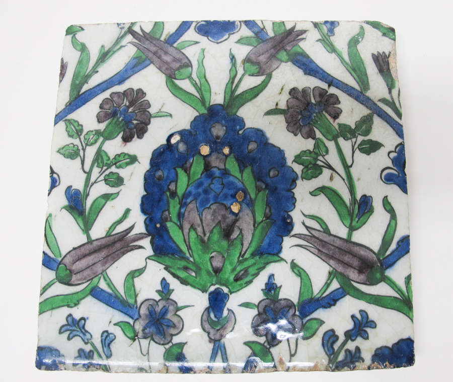 White square tile painted with a dense symmetrical floral pattern in blues, blacks and greens. A large maned flower occupies the center, with smaller flowers of varying shapes surrounding it.
