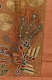Close-up of a detailed red, green, blue, and gold embroidered bird with peacock feathers surrounded by swirling clouds and gold discs on an orange silk background.