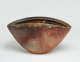 A deep clam shaped stone bowl with a wide opening and a gray-green rim. Shell like etchings outline the outer bowl which is dark brown on the left and a rich red brown on the right.