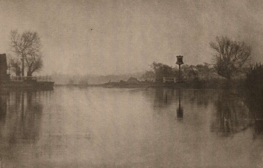 A black and white image depicting a calm river bordered by bare trees on each side with a small structure on the right.