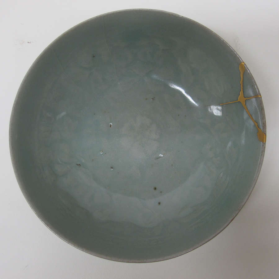 Top-view of a muted teal glazed bowl’s interior with faded floral patterns and small gold patched cracks on the right top edge. 