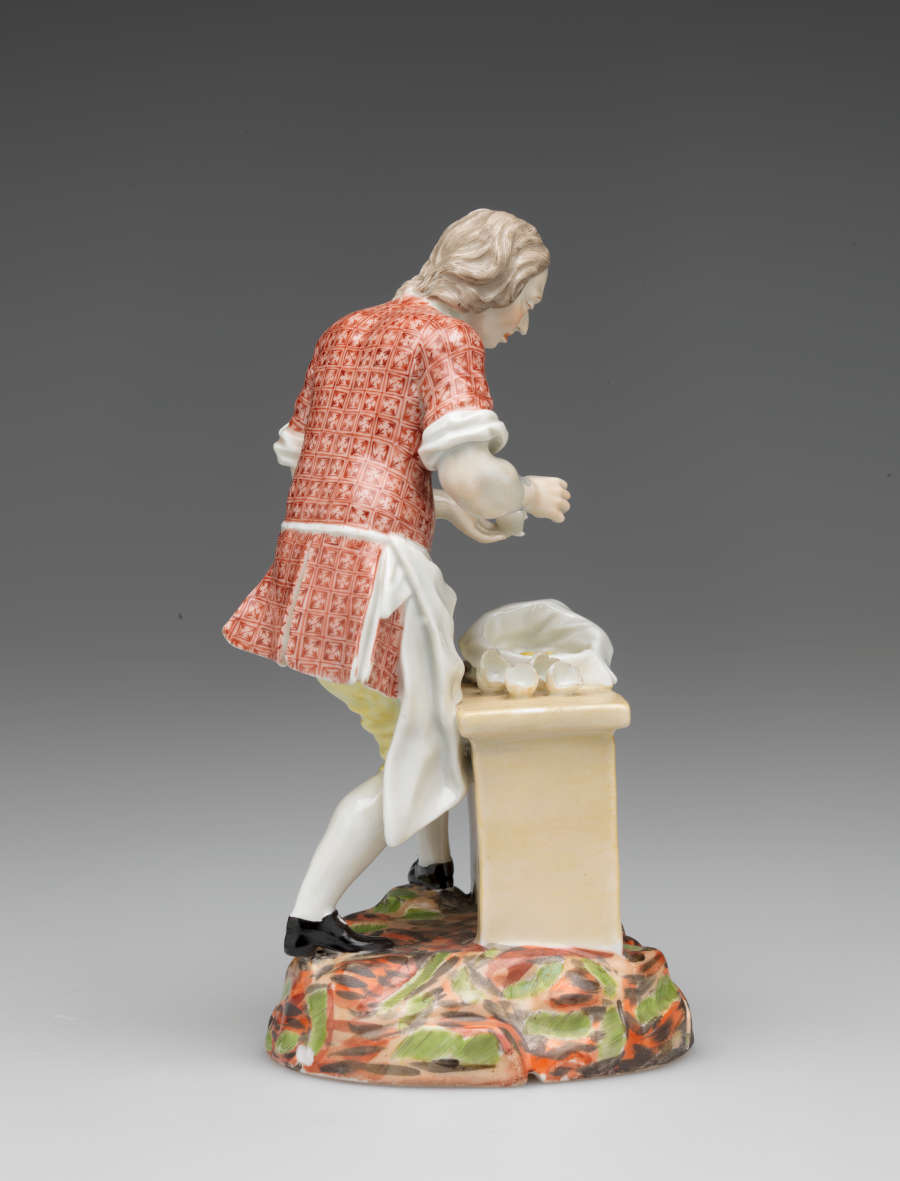 A sculpture of a figure in historical clothing and a table, a sack, and broken egg shells. The ground is brightly colored red, brown, and green.