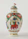 A tea caddy with a lid and green, red, blue, pink, yellow and gilded decorations. The finial is a sculptural flower white, red and green.