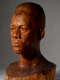 Three quarter view of a dramatically lit, glossy wooden bust depicting the head of a male figure with afrocentric facial features. He has a stern expression and an intense gaze.